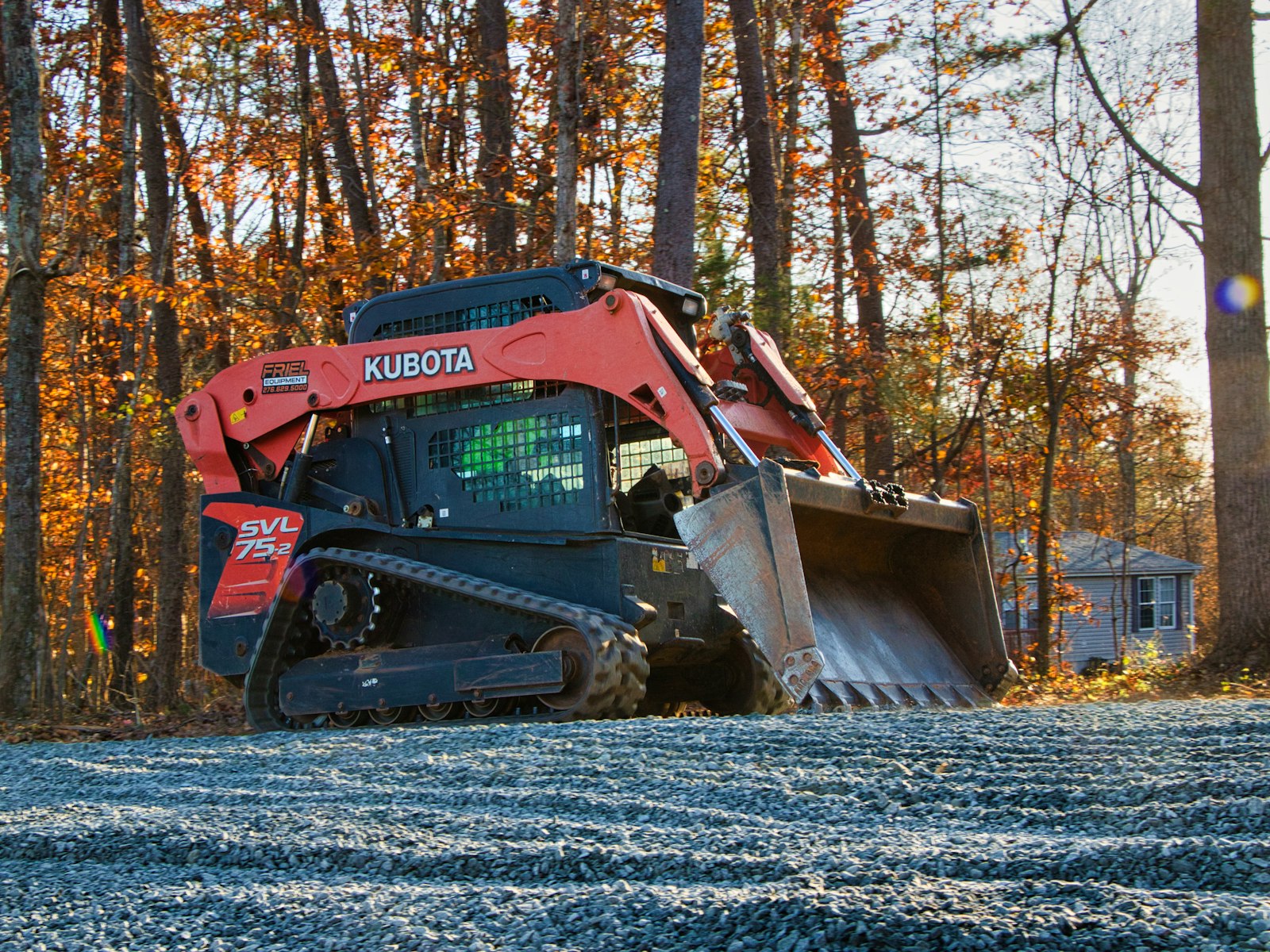 Orange compact track loader with bucket on a bed of crushed gravel beside a residential house