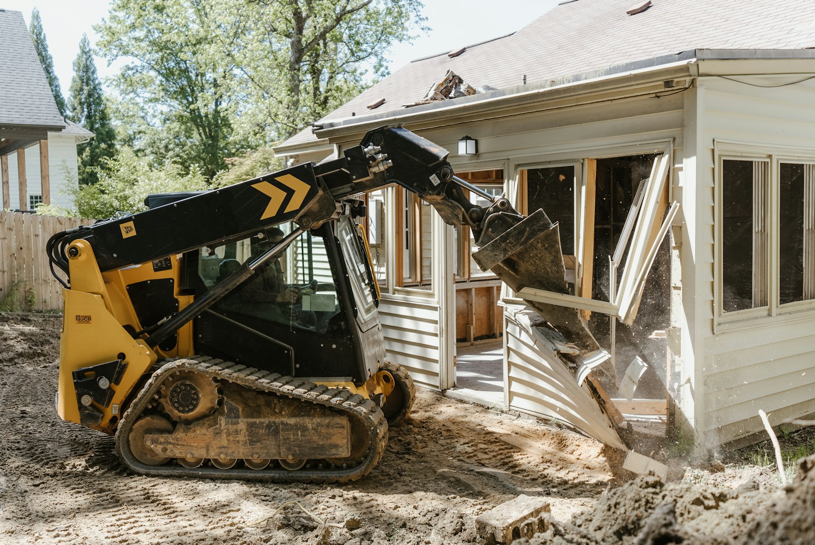 Compact track loader clearing a residential addition and churning dirt in a backyard near a wood fence